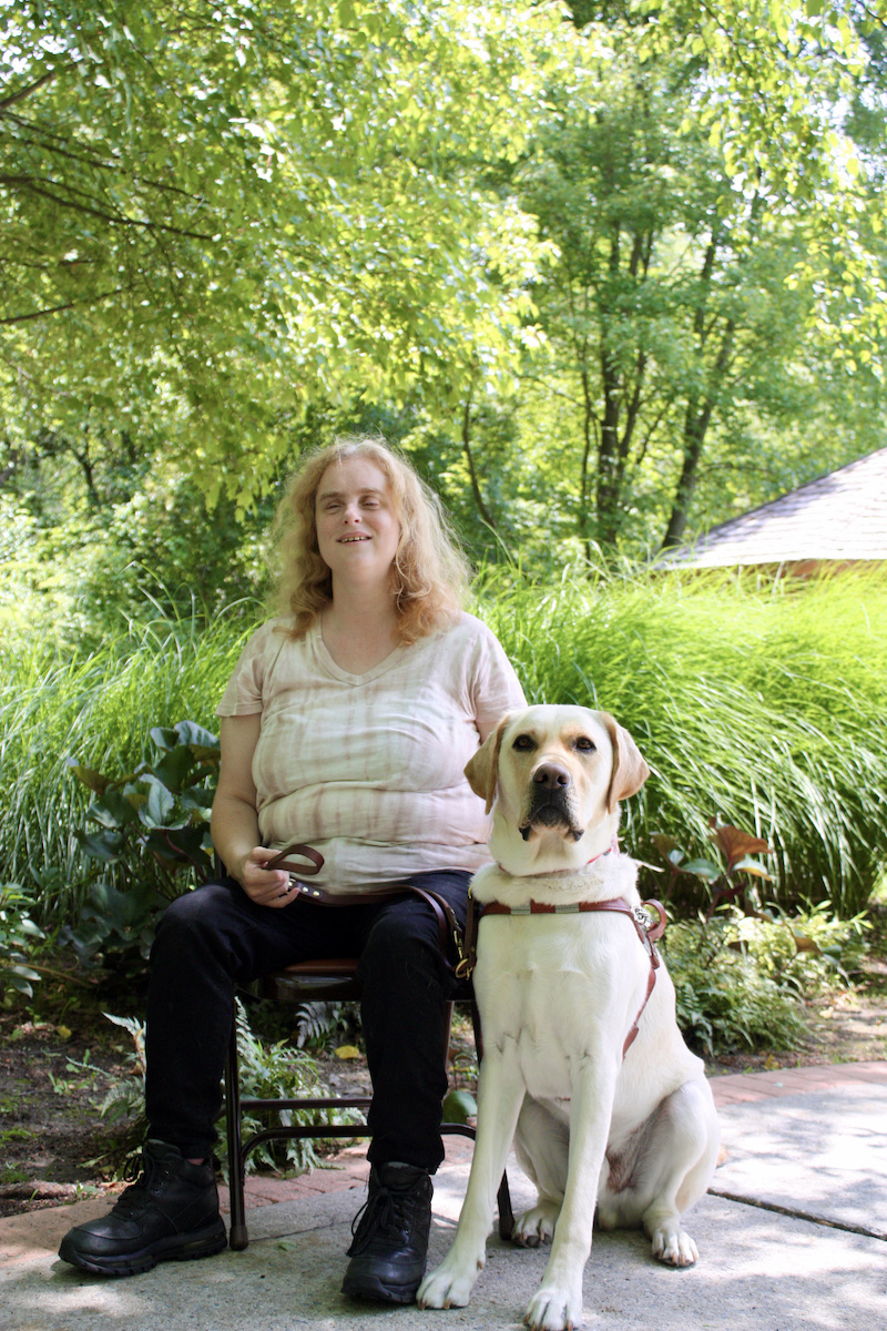 Graduate Sarah and guide dog Windsor against a lush green summer background