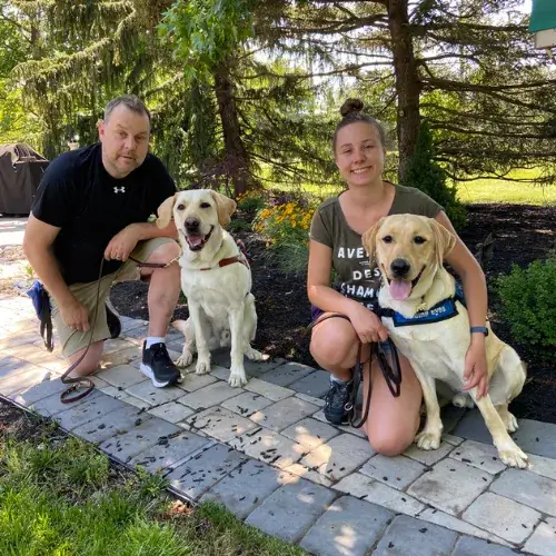 Yfke kneels on the stone path with yellow lab pup on program Lancelot in a sit by her side