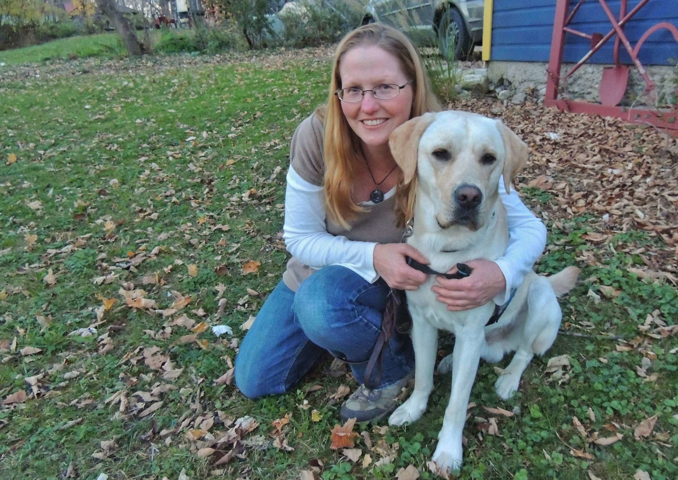 Tara kneels in the grass with her arms around a sitting yellow lab named Nacho.