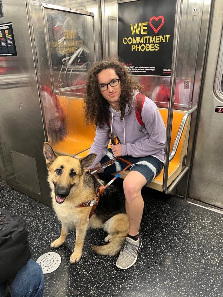 Kai and his guide dog pride, a female black and tan German Shepherd in harness, both sit on a New York City subway. Pride sits between Kai’s legs as she calmly smiles towards the camera