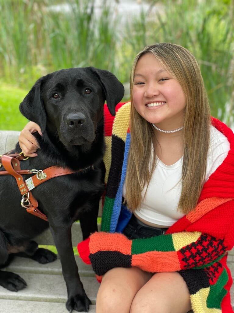 Graduate Sydney and her guide dog Vaughn, a black male lab in harness, both sit on a bench on the nature path bench at Guiding Eyes for the Blind training school campus with the grassy pond in the background