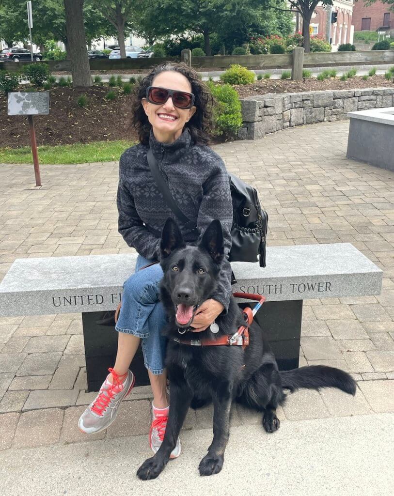 Nicole and her guide dog Hedwig, a female black German Shepherd in harness. Nicole sits on the granite bench, smiling in her sunglasses and gently lays her hand over Hedwig’s shoulder as sits calmly by her feet.  Nicole lays her arm over Hedwig’s shoulder as he sits so closely to her lap