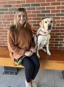 Graduate Carly and yellow Lab guide dog Sharon sit on a bench against a brick wall