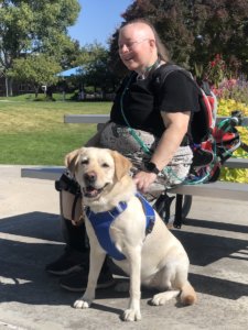 Doula sits on a sunny bench with smiling yellow lab guide dog Bushka
