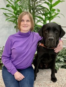 Graduate Kira and black Lab guide dog Ferrari sit in front of a tall potted plant