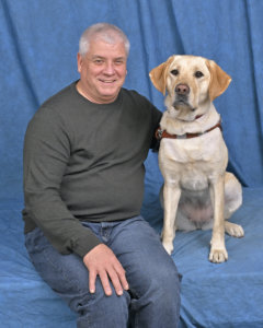 graduate Paul sits next to yellow Lab guide dog Collin against a blue background