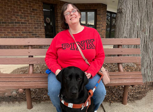 Amanda sits on a wood bench and black Lab guide Brava at her feet