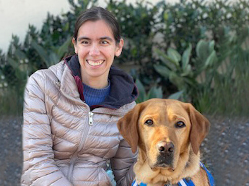 Miriam and yellow lab guide dog Yani sit against green foliage for team portrait