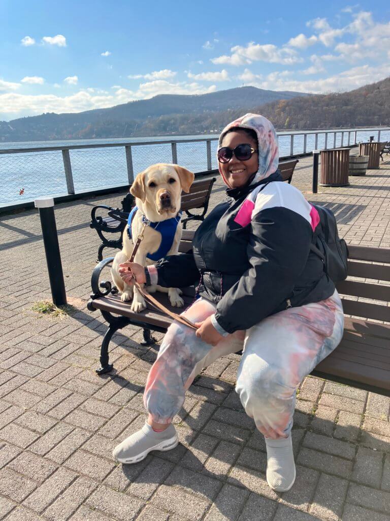 Briasa sits on a bench by the Hudson with guide dog Journey