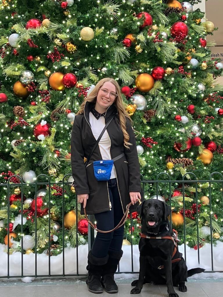 Michaela stands in front of a brightly decorated tree while guide Eleanor sits at her feet with tongue out