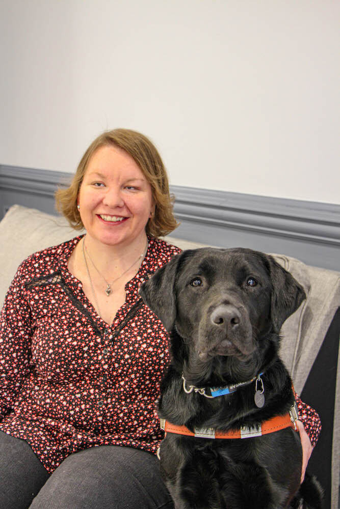 Caitlin sits in upholstered chair with black Lab guide Gemini for team portrait