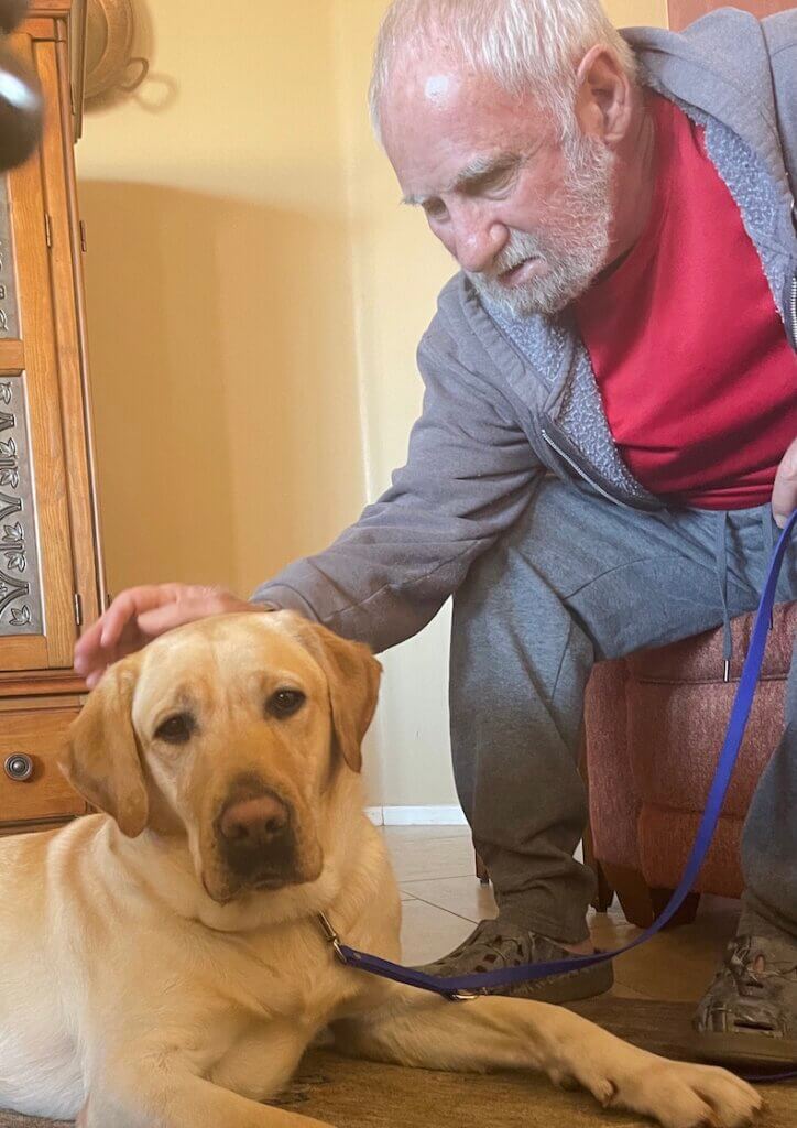 Dick leans forward from his chair to pat guide dog Presley on the head