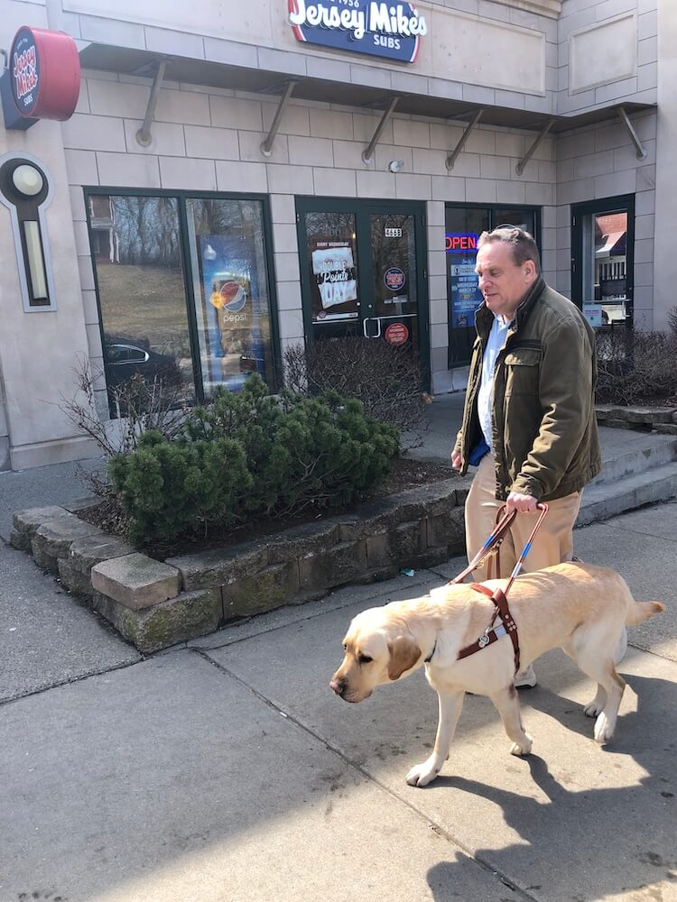 Carl and Tigger walk quickly and safely down a sidewalk past a sub shop
