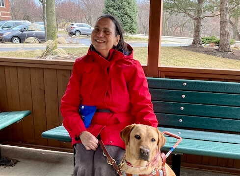 Cindy and yellow lab guide Barlie sit on green bench in park-like setting