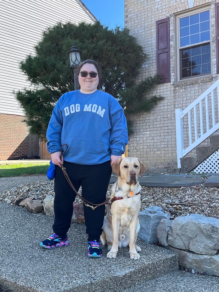 Jessica stands in a blue Dog Mom shirt with yellow Lab guide dog Uber