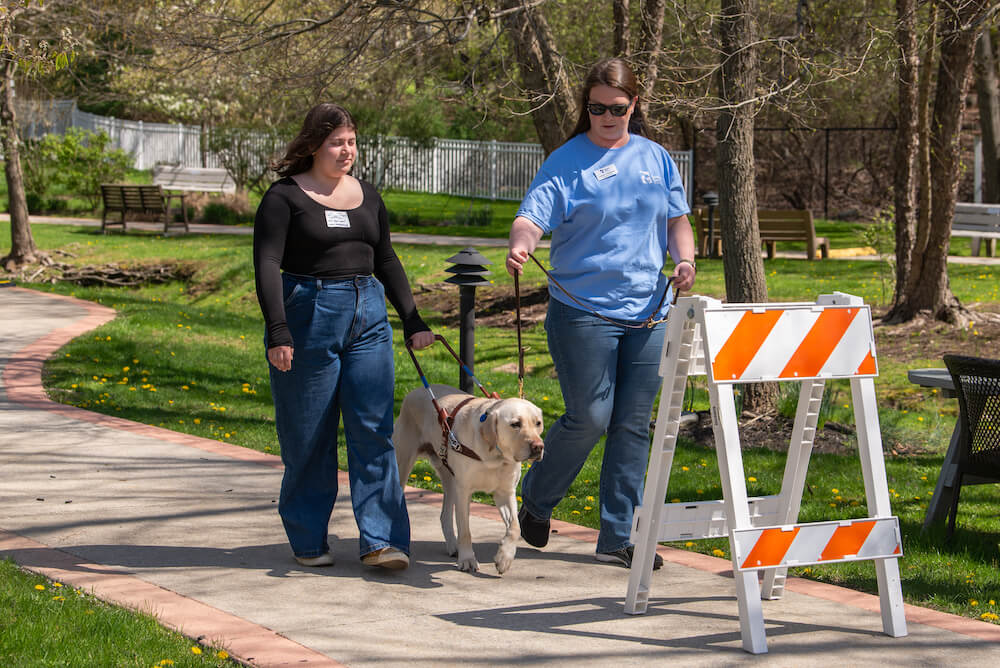 a volunteer blindfolded walks with a dog in harness with a trainer holding support leash