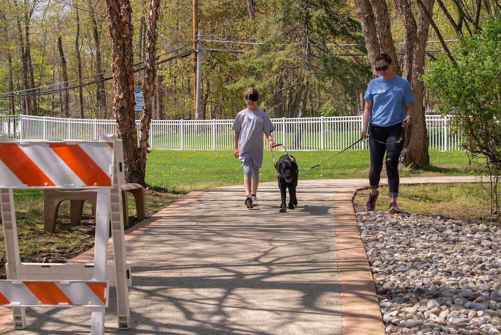 a boy in blindfold walks with a black dog in harness with a trainer holding support leash