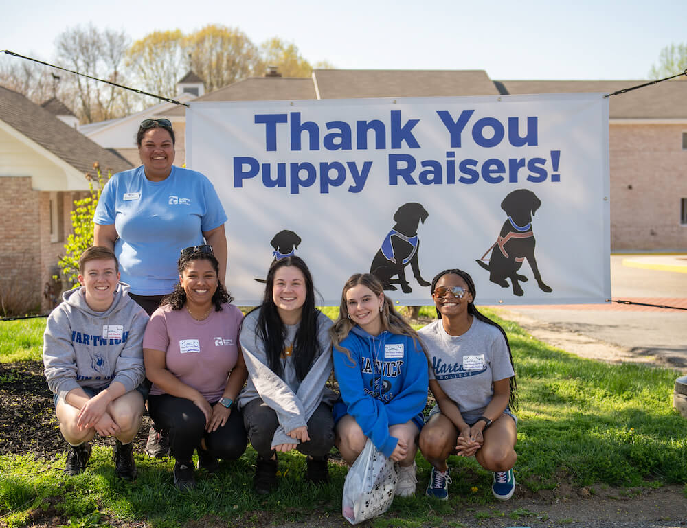 Puppy raiser kneel in front of banner sayin Thank You Puppy Raisers!