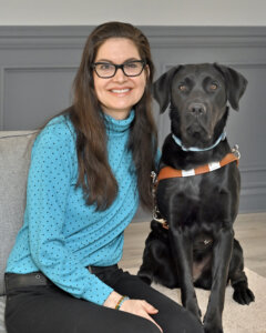 Wendy smiles sitting next to black Lab guide Charlene on a gray bench for team portrait