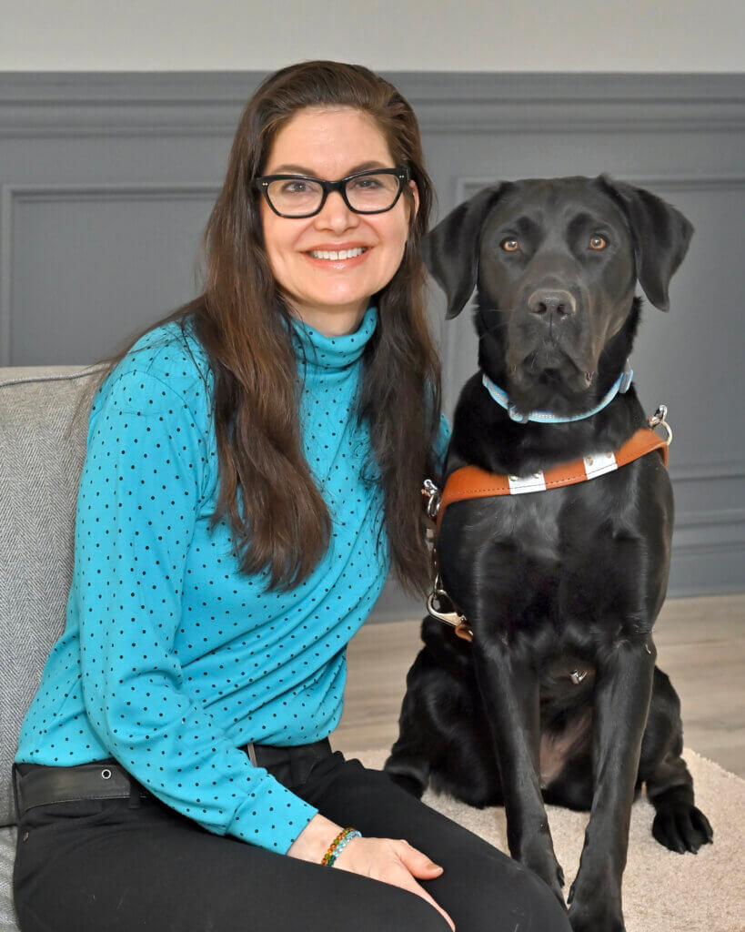 Wendy smiles sitting next to black Lab guide Charlene on a gray bench for team portrait