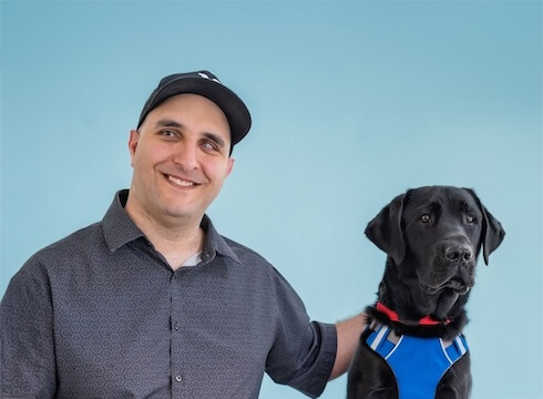 Aziz and black Lab Guide Dog Dundee pose for their graduate portrait in front of a light blue background