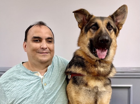 Carlos and German Shepherd guided dog Indo sit indoors for their team portrait