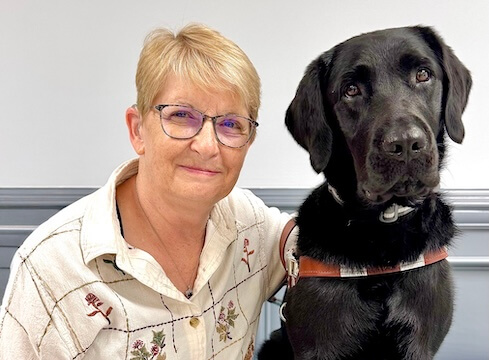 Connie sits beside black Lab guide dog Rhonda against a gray background for their team portrait