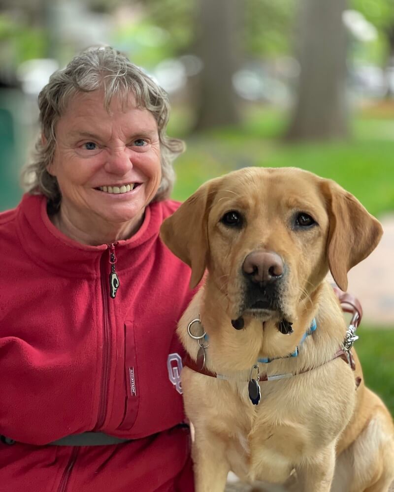 Darla in red and yellow guide dog Biscotti sit side by side outdoors for team portrait