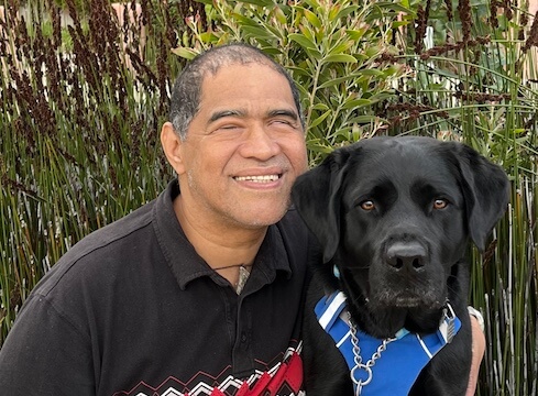 Emmanuel sits close to black Lab guide dog Arby in an outdoor setting