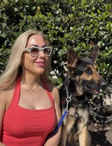 Irma sits with German Shepherd guide dog Wonder in a bright sunny location in front of green foliage