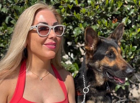 Irma sits with German Shepherd guide dog Wonder as they both look to their left in a bright sunny location in front of green foliage