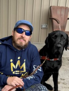 At a residential entrance, Jason squats down next to black Lab guide dog Yodel as they look to the camera. 