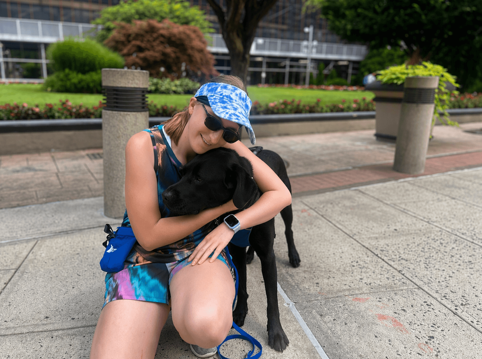 Maryann kneels on a paved area in front of a building with her arms lovingly around guide Dusty