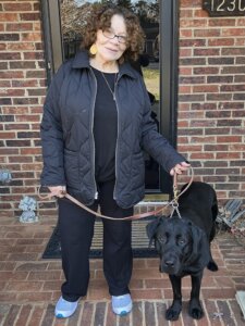 Sandra and black Lab guide dog Elliott stand at entrance of a brick building looking at the camera