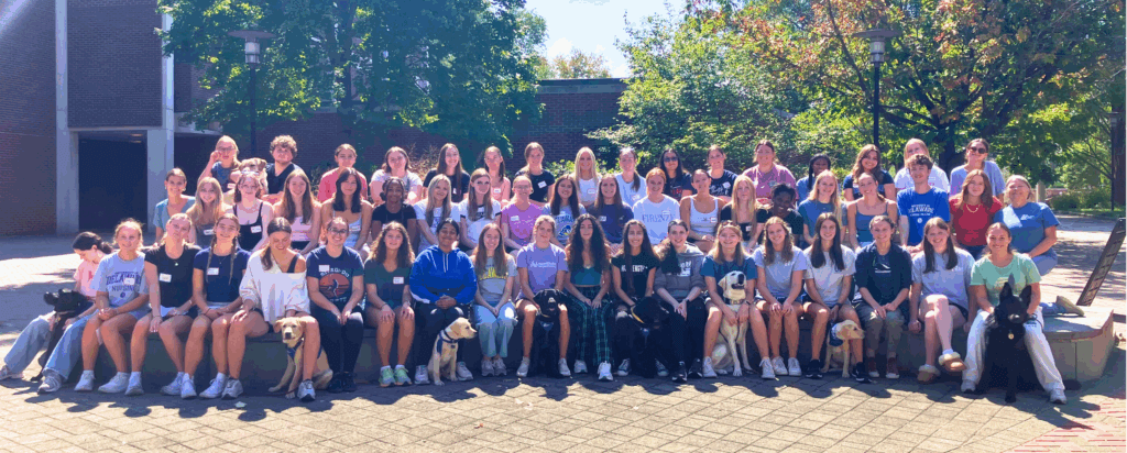 Large group of students gather in a courtyard of University of Delaware and several Guiding eyes pups sit at the front row