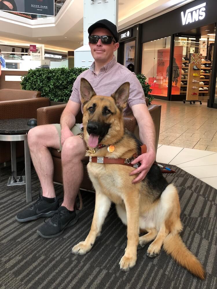 Zachary sits in a chair at a shopping mall with arm around German Shepherd Fritz