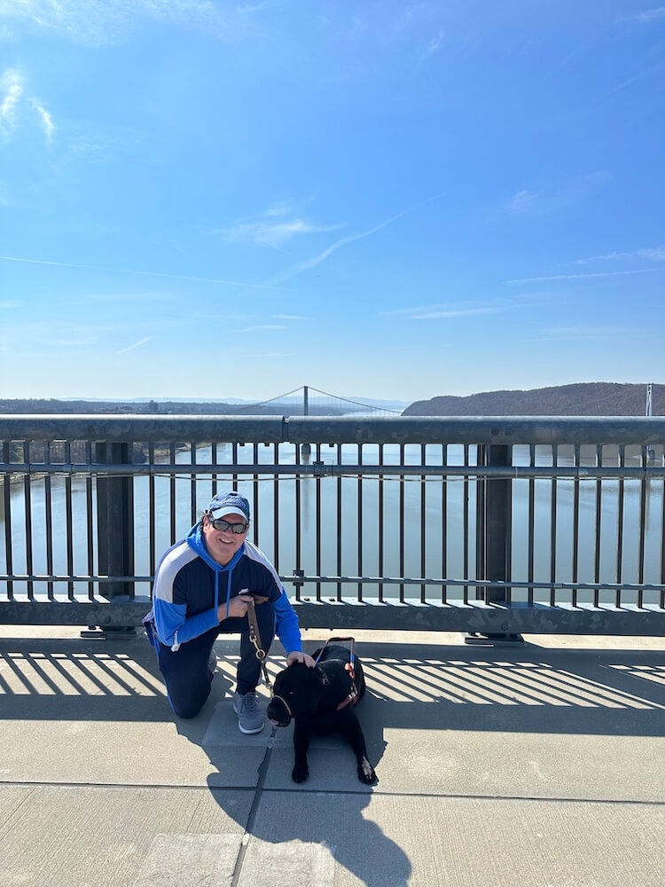 Christian kneels next black lab guide Gideon on the Walkway bridge over Hudson River