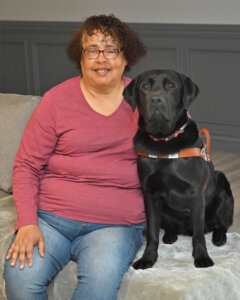 Jane sits close to black lab guide dog Noah inside on a covered bench for team portrait
