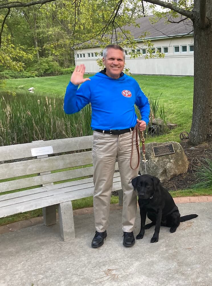 Joe waves to the camera as he and Martha stand near a bench on campus
