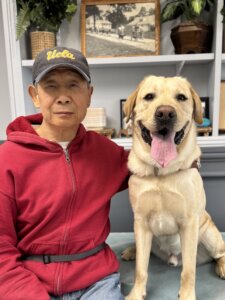 Mark and yellow Lab guide dog Justice sit in front of shelves filled with various items for their team portrait