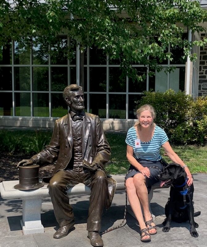 pup on program Neptune sits with raiser Anne and a Lincoln statue sitting on a bench