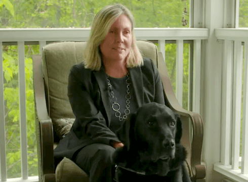 Karen sits with black lab guide dog Flint on porch with wood railings