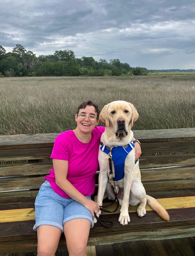 Kandi and yellow Lab guide dog Yacht sit on a bench with fields behind them