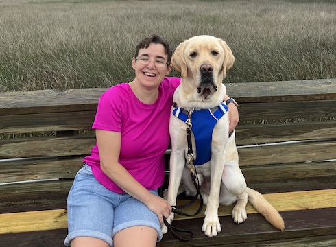 Kandi and yellow Lab guide dog Yacht sit on a bench with fields behind them
