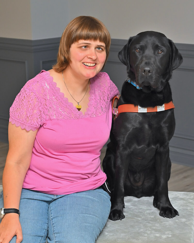 Sarah sits with black Lab guide Sully for grad team portrait