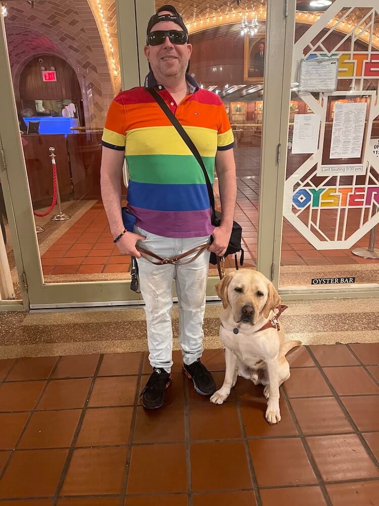 In NYC Gabriel stands at storefront in bright rainbow striped shirt with Gabe sitting below
