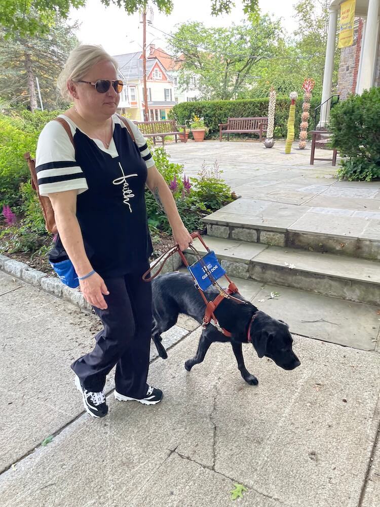 Mandy and black lab Reina walk along pavement avoiding obstacles to their left
