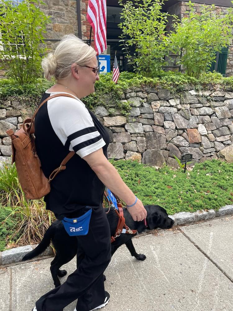 Mandy and black lab Reina walk along pavement with a raised curb to grassy area
