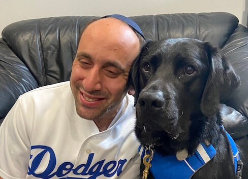Menachem in a Dodgers shirt leans in towards black Lab guide dog Sumatra