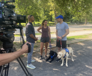 Interviewer David Scott stands in the park with Payden, Thomas and two guide dogs with a filming camera in foreground